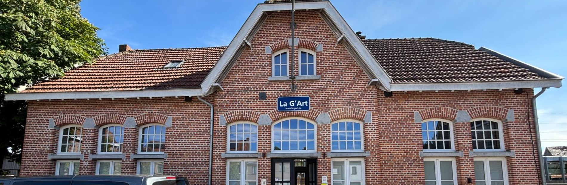 Vooraanzicht van een roodstenen gebouw met een bord "La G'Art" en een geparkeerde grijze bestelwagen.